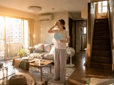 日差しの強いリビングで、エアコンと扇風機を併用しながら汗を拭う暑そうな女性